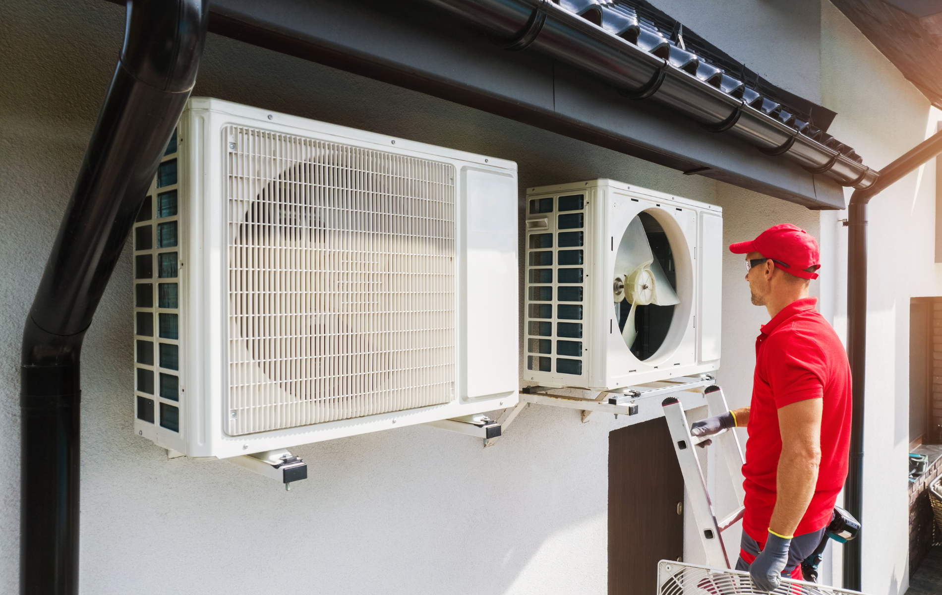 HVAC technician in red shirt and cap works on white air conditioning units mounted on a building.
