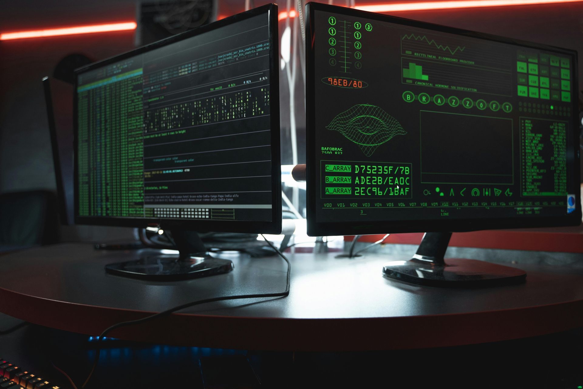 Two computer monitors displaying green code on a dark desk in a dimly lit workspace