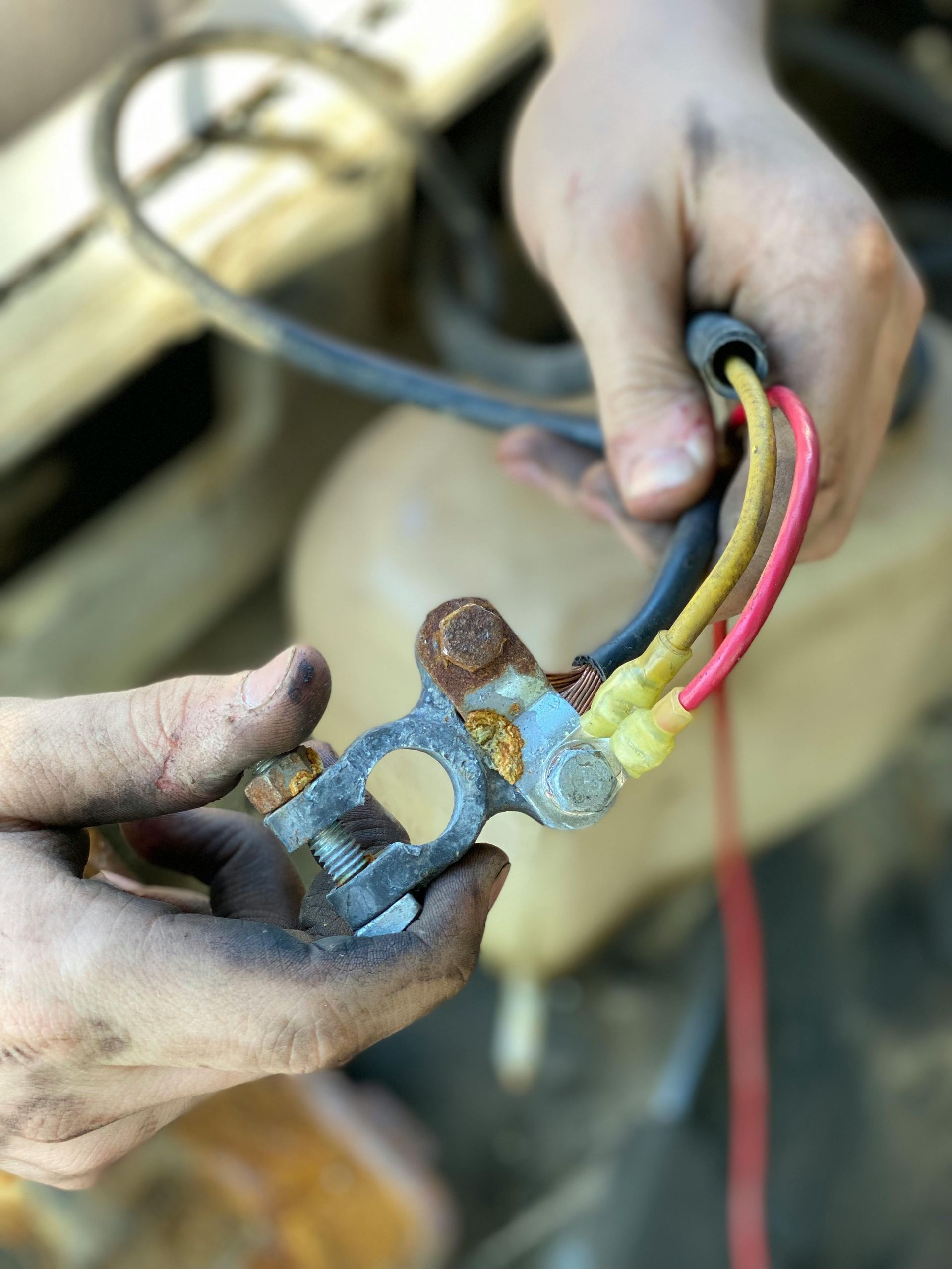 Hands holding a corroded car battery terminal with red, yellow, and black wires, in an engine bay.