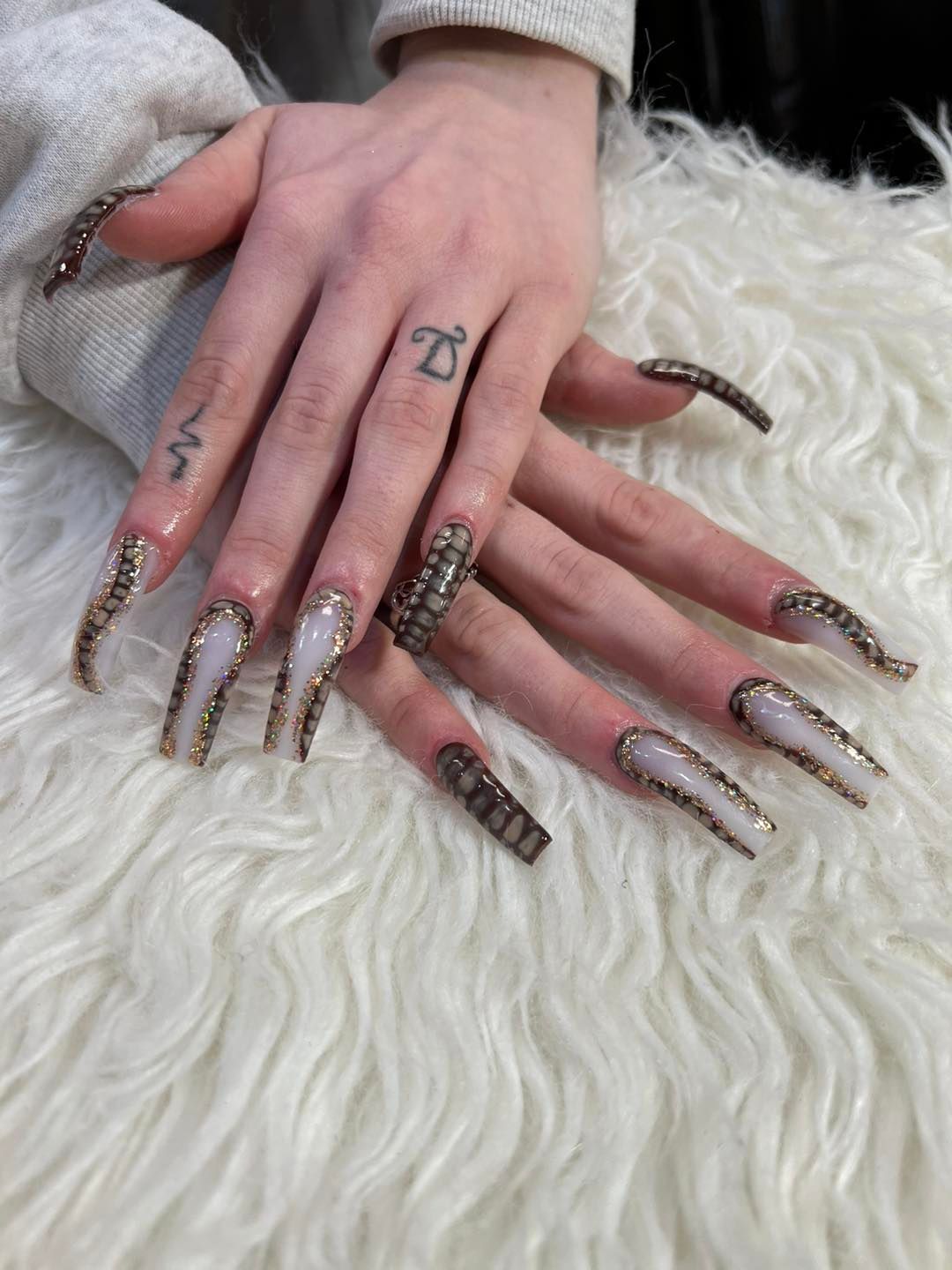 A pair of hands with long, square-shaped acrylic nails featuring a white base with intricate dark brown snake-print designs.