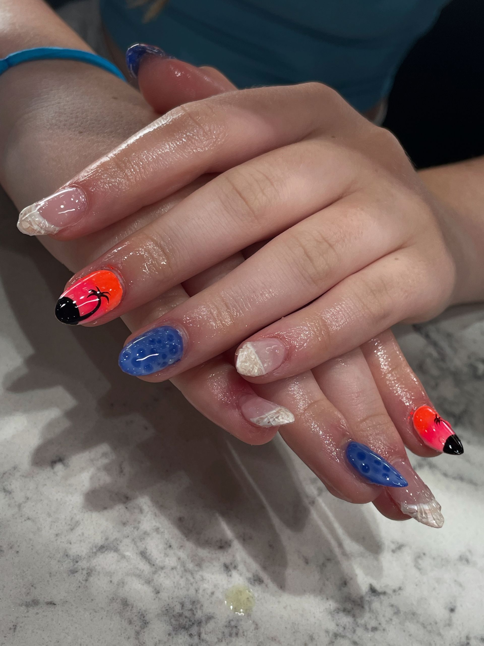 Manicured hands with alternating glitter, solid blue, and sunset-style gradient nails featuring palm tree silhouettes.