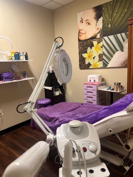 A spa treatment room featuring a facial machine, a magnifying lamp, a reclining bed with purple linens, and wall decor.