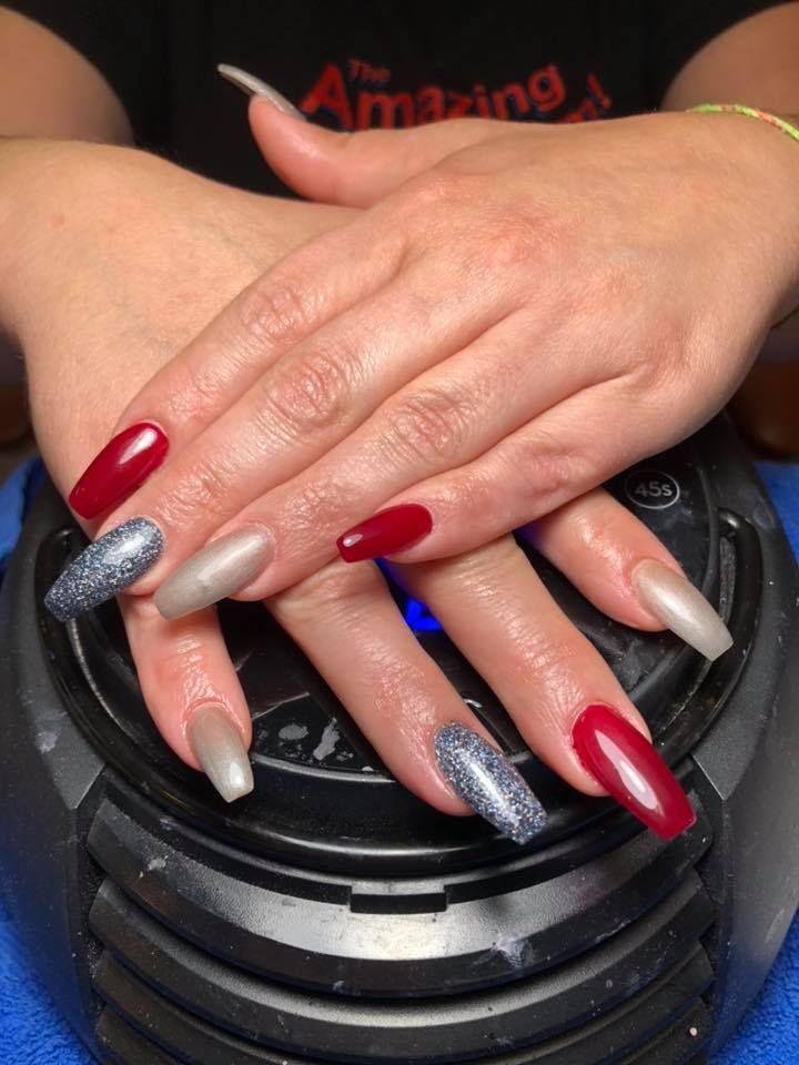 Hands with coffin-shaped nails painted in red, silver glitter, and nude polish resting on a black nail lamp.