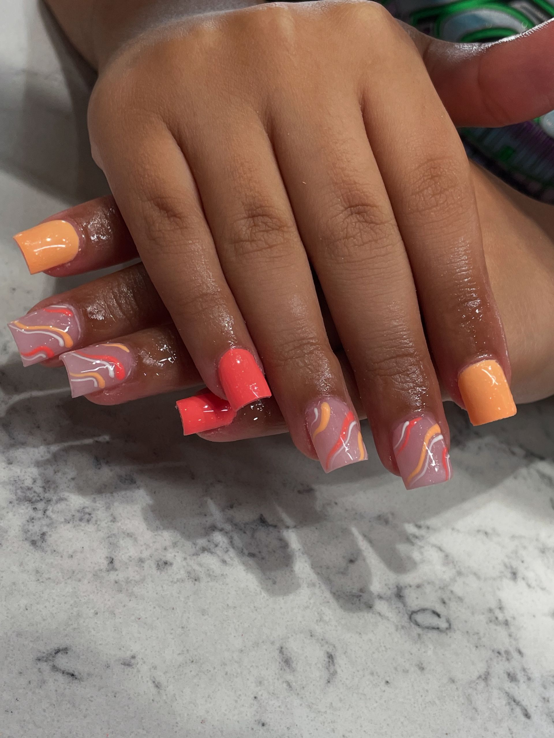 Hands with square-shaped nails painted in alternating solid coral and sheer base colors with orange and white swirl art.