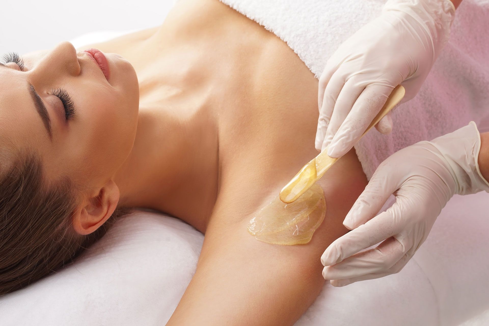 A person receiving professional underarm waxing with a wooden applicator by an aesthetician wearing gloves.