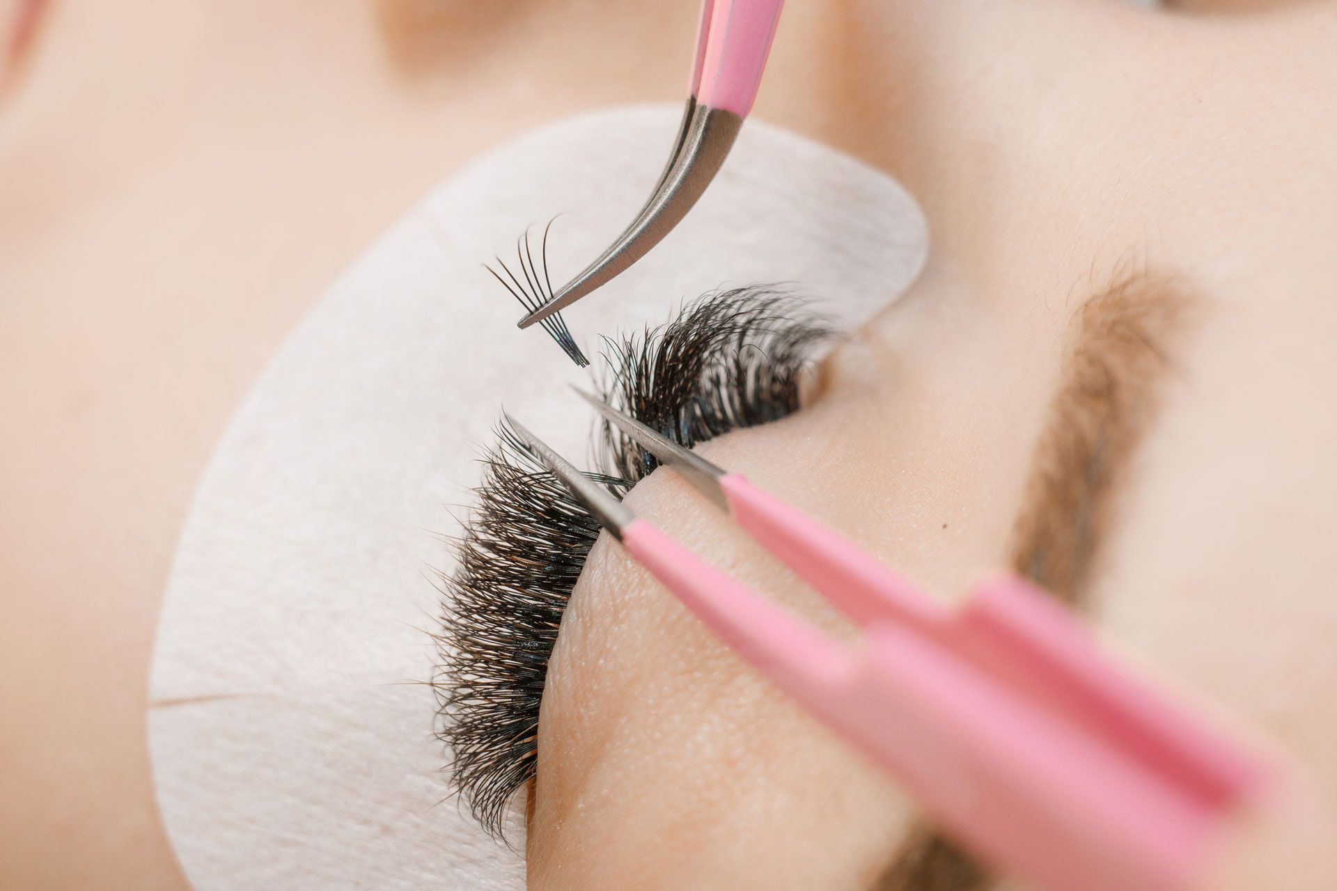 A technician uses pink tweezers to apply a volume eyelash extension fan to a person's natural lash over a white eye pad.