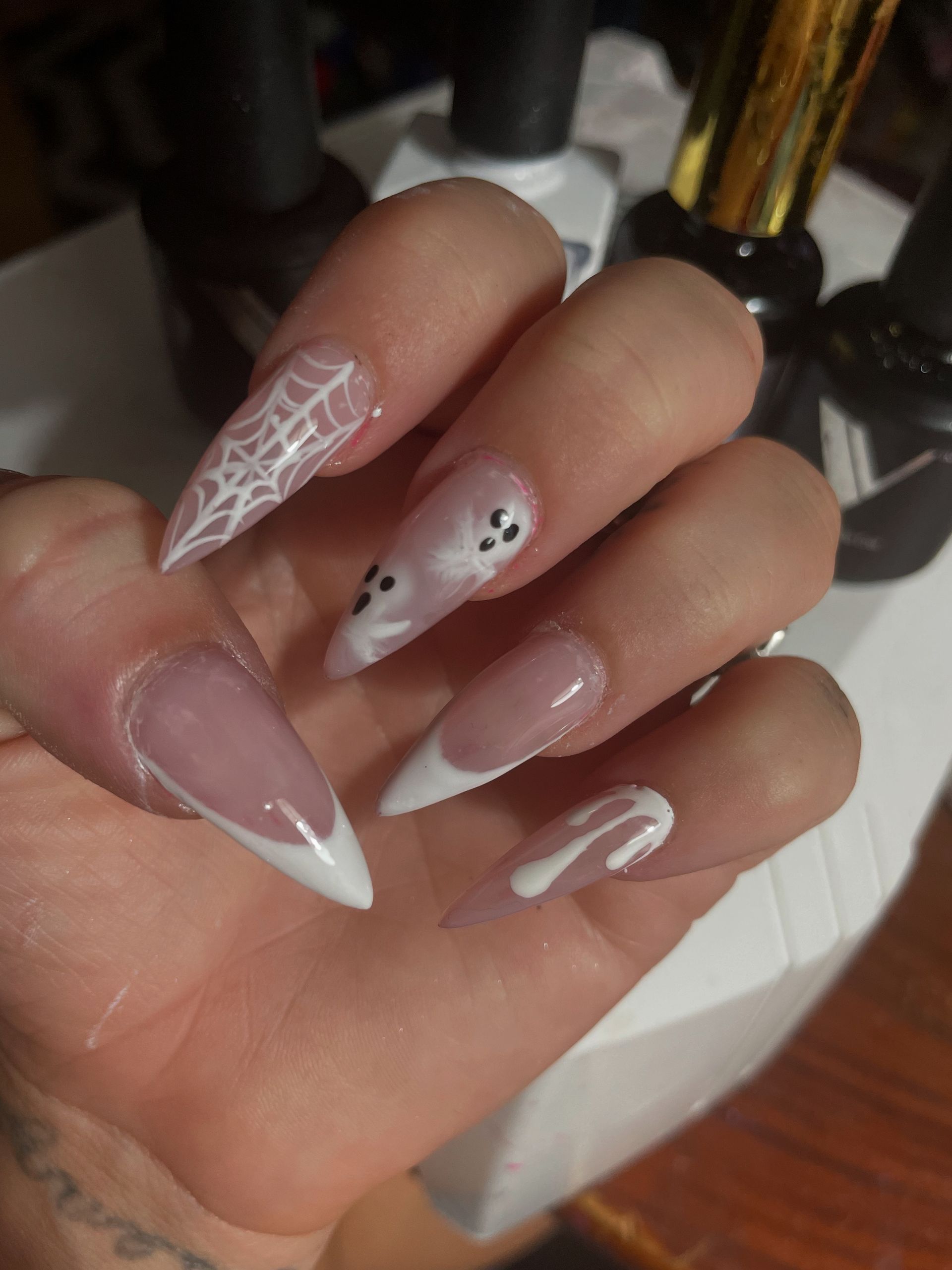 A hand with long, almond-shaped nails featuring various white Halloween-themed designs: spider webs, ghosts, and drips.
