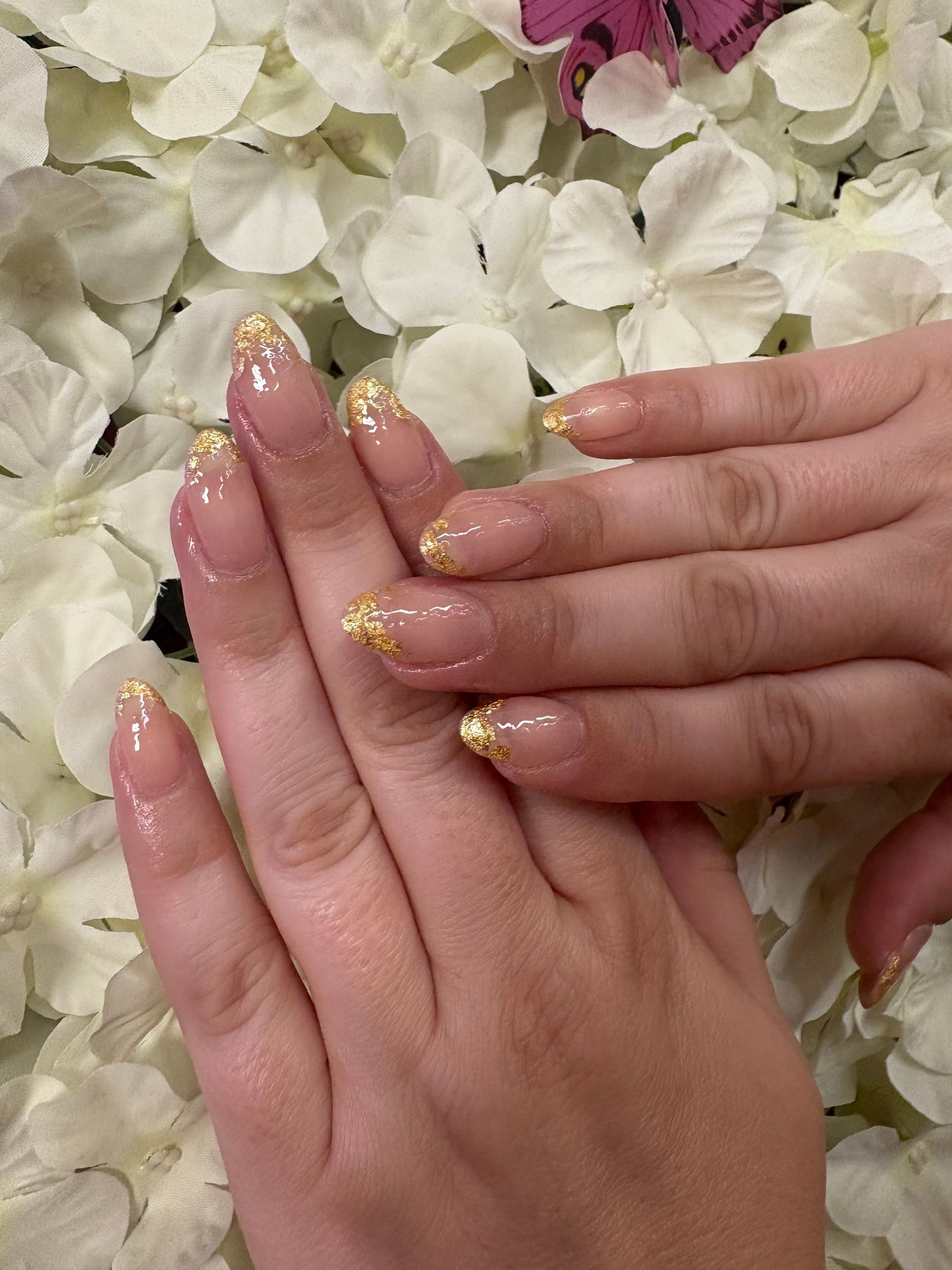 A close-up of hands with manicured nails featuring a sheer nude base and gold leaf tips, posed against white flowers.