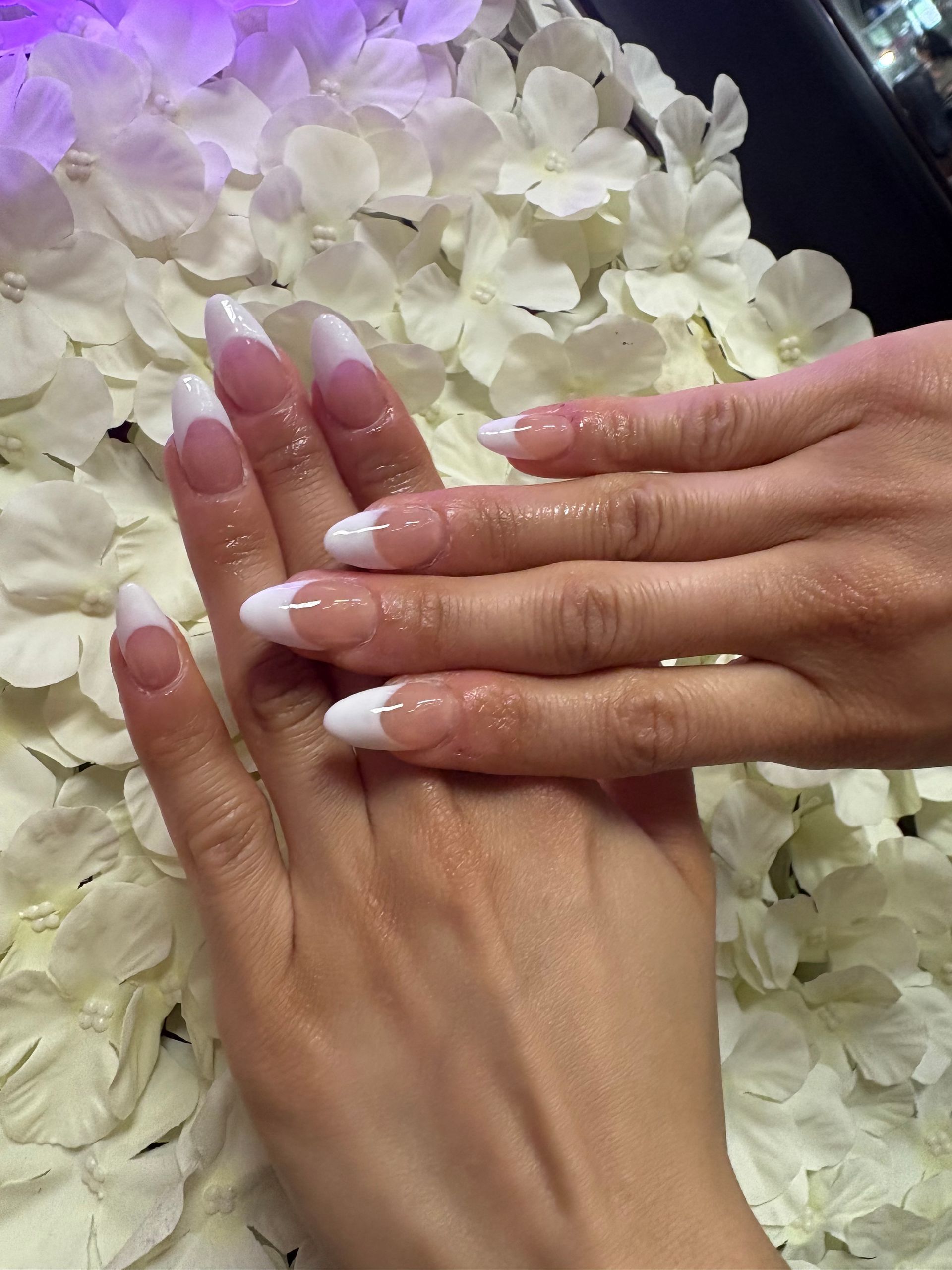Hands with almond-shaped French manicure nails with gold glitter accents, positioned over white flower petals.