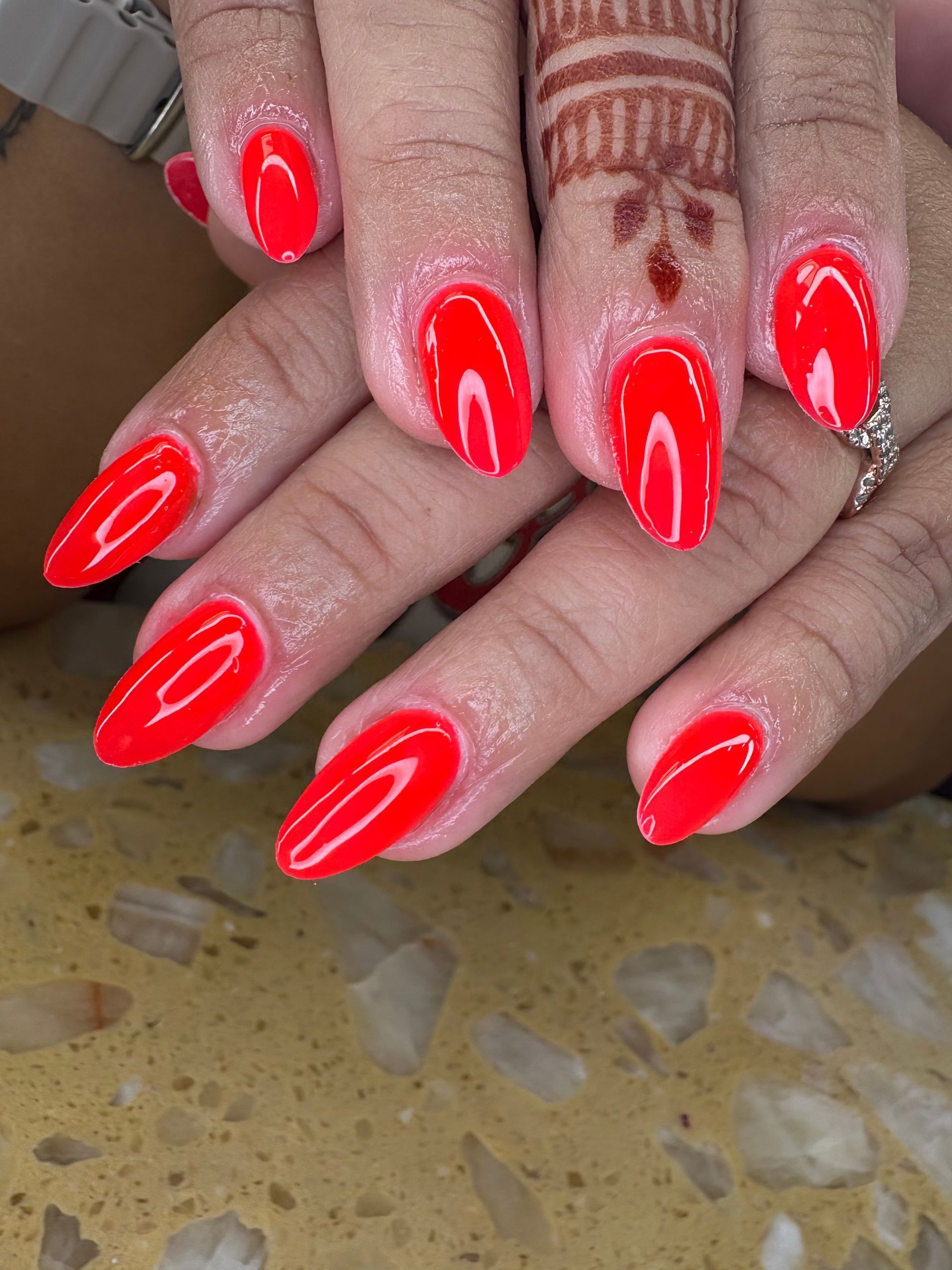 Close-up of hands with neatly manicured, almond-shaped bright red nails, one finger features a delicate henna pattern.