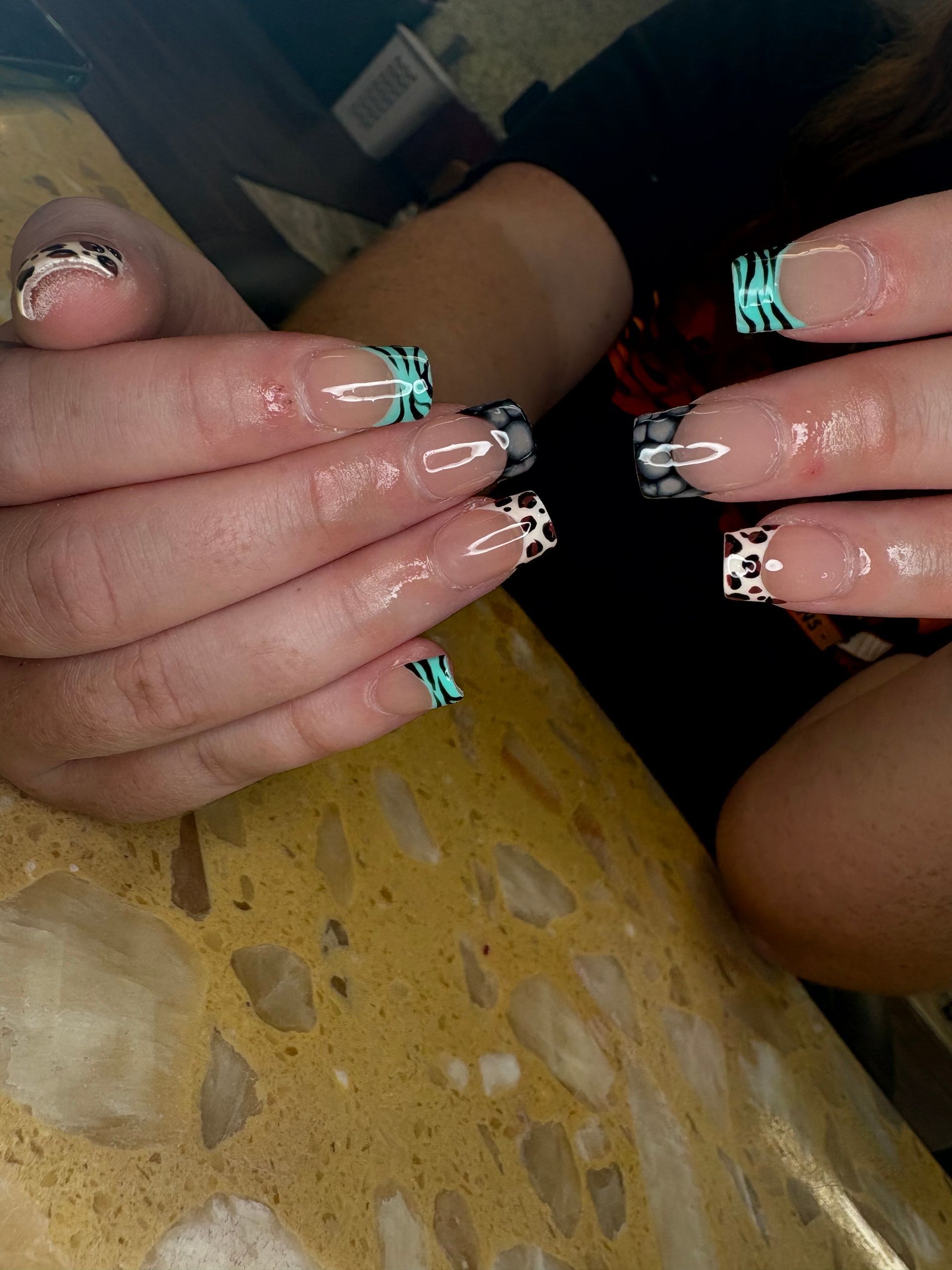 Hands displaying long, square-tipped nails featuring a natural base with colorful animal print and zebra stripe tips.