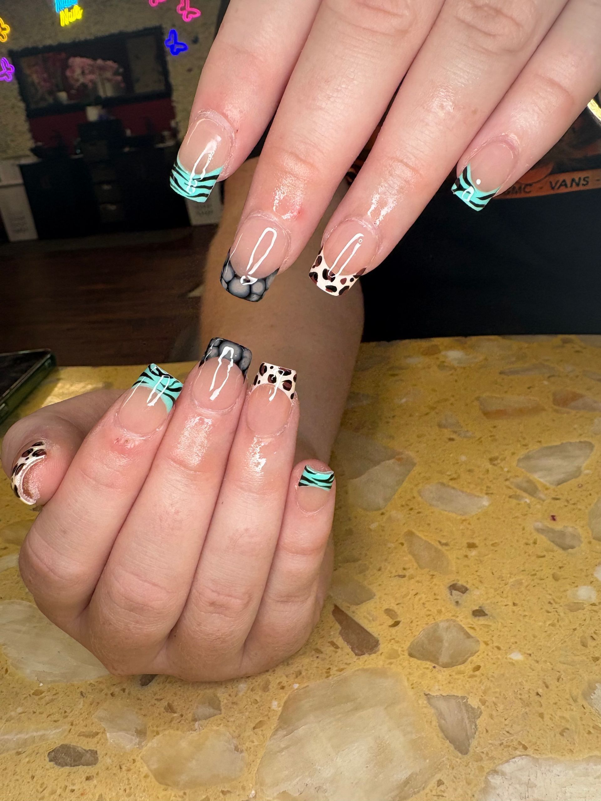 A close-up of hands showing French-tip nails with alternating teal zebra print and black and white leopard print patterns.
