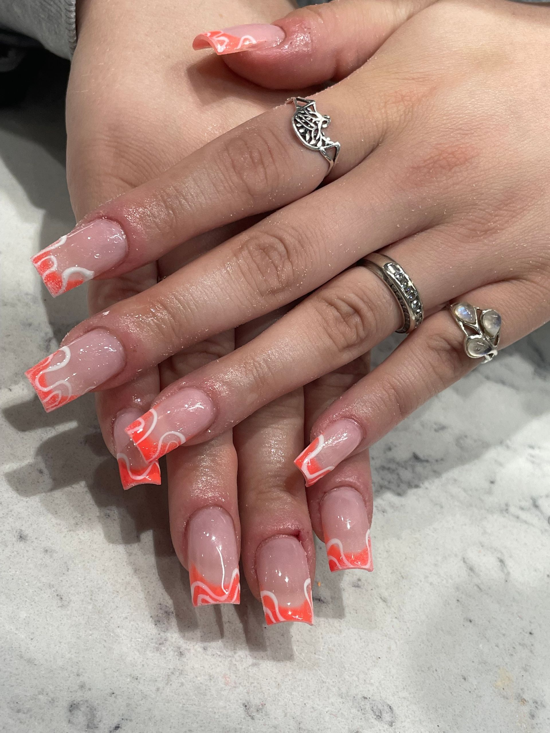 Square-shaped nails with a clear base and orange-and-white swirling tip designs, featuring three silver rings on fingers.