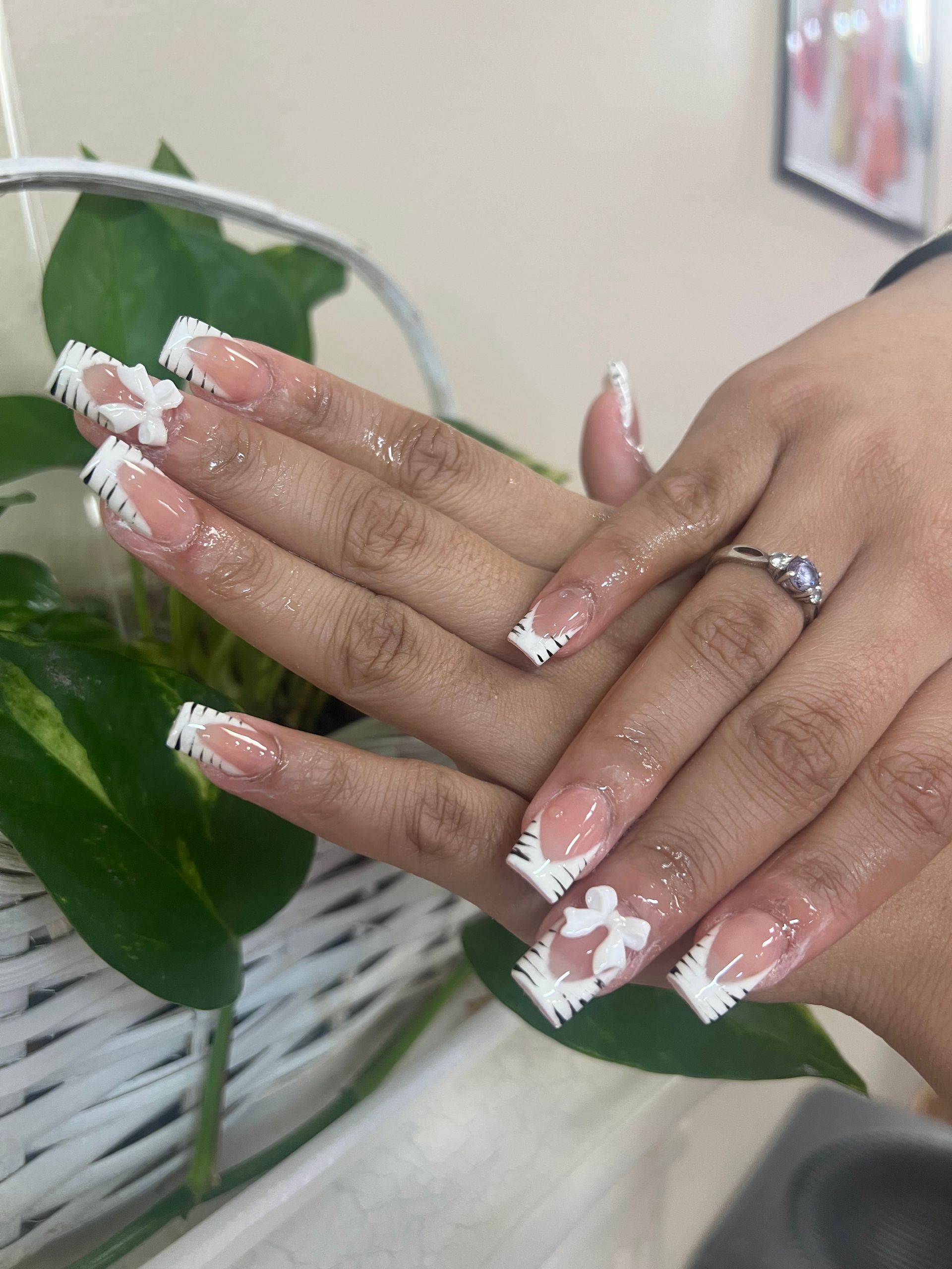 Hands displaying long, square-tipped nails featuring a white French manicure and 3D white bows against a green plant.