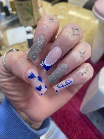 A hand with long, square-shaped acrylic nails featuring blue heart patterns, glitter, and white lace-style French tips.
