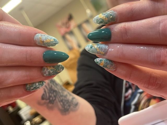 Hands displaying a manicure with alternating teal solid and teal-and-white marble nails decorated with gold flakes.
