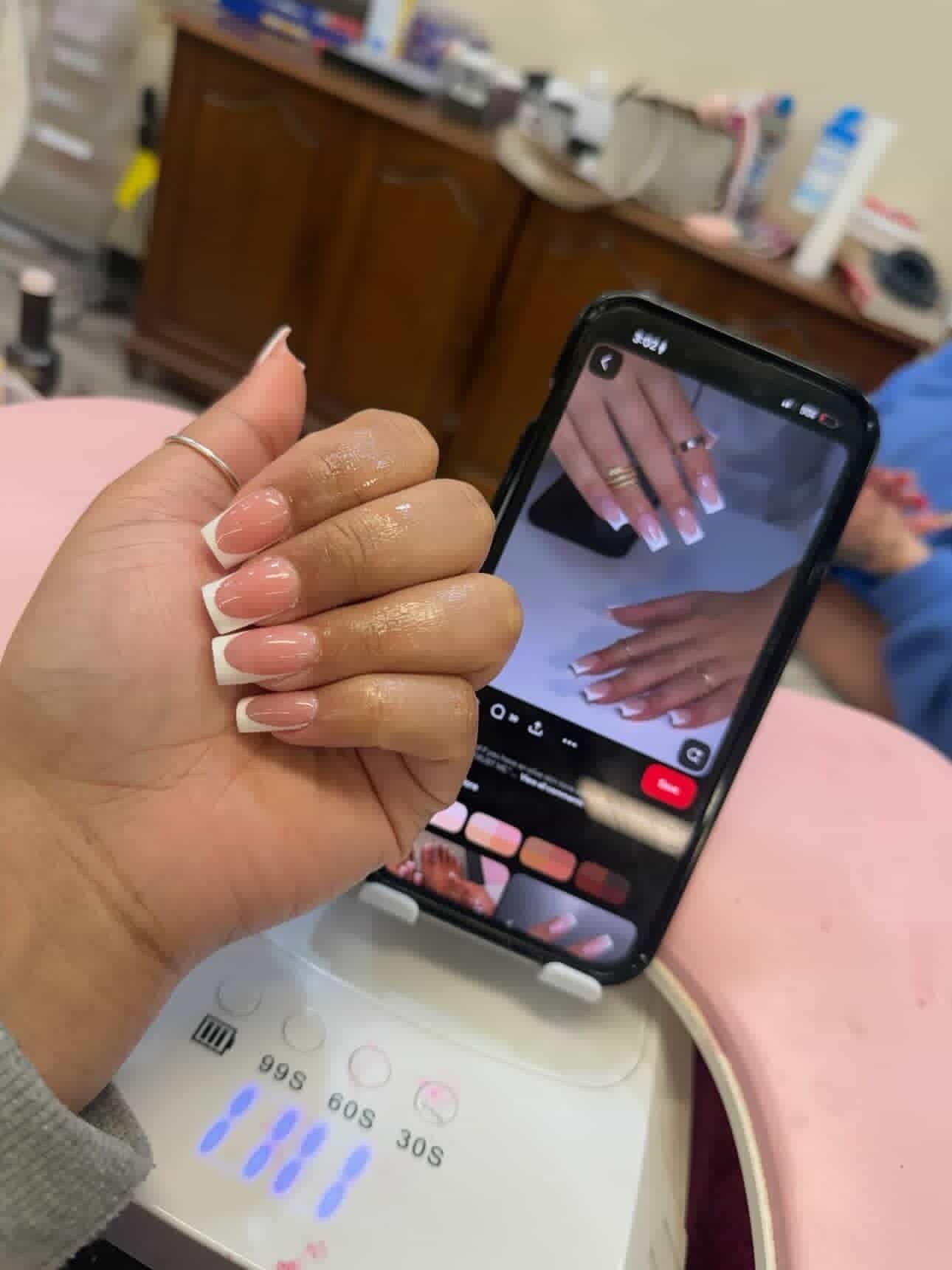 A hand displays a fresh French manicure next to a smartphone showing a reference photo of the same nail style.