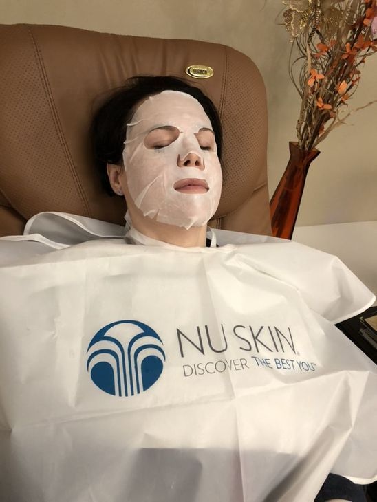 A person lying in a chair wearing a white facial sheet mask with a Nu Skin branded cape.