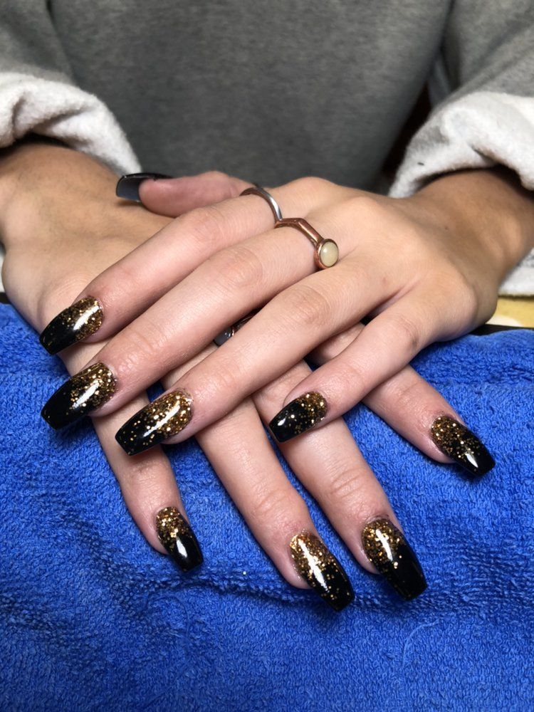 Hands with long, square-shaped nails painted black with a gold glitter ombre fade, resting on a blue textured cloth.
