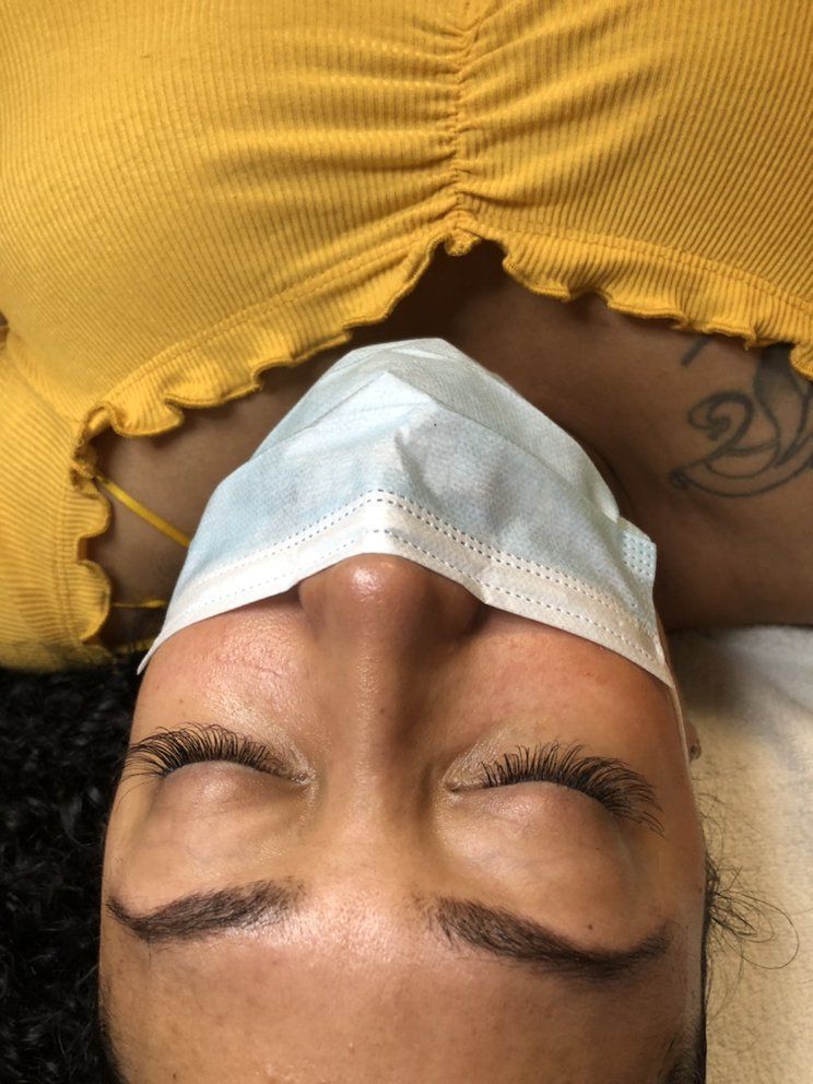 A person wearing a light blue face mask lies on their back, their eyes closed with dark, defined eyelashes.