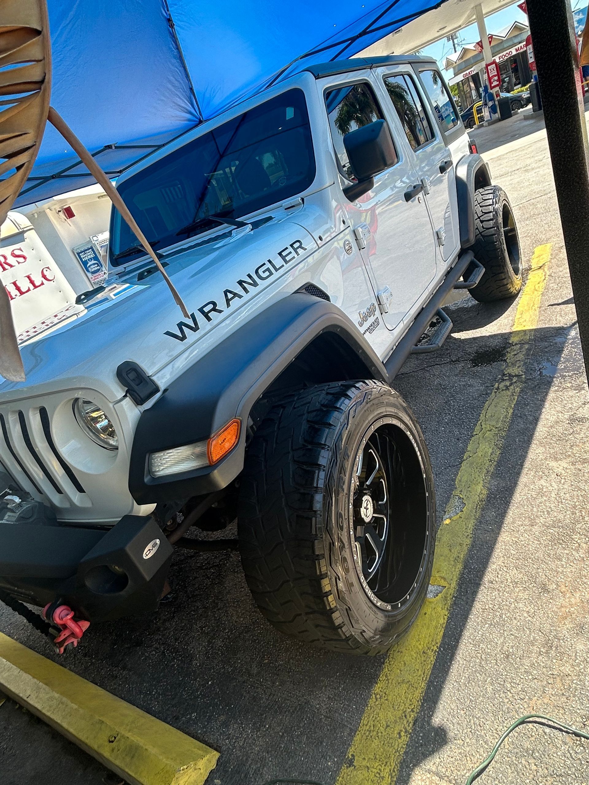 White Jeep Wrangler with large black tires parked outside, under a blue awning.