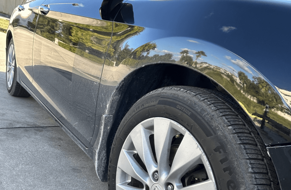 Black car's front tire and fender, reflecting a sunny outdoor scene.