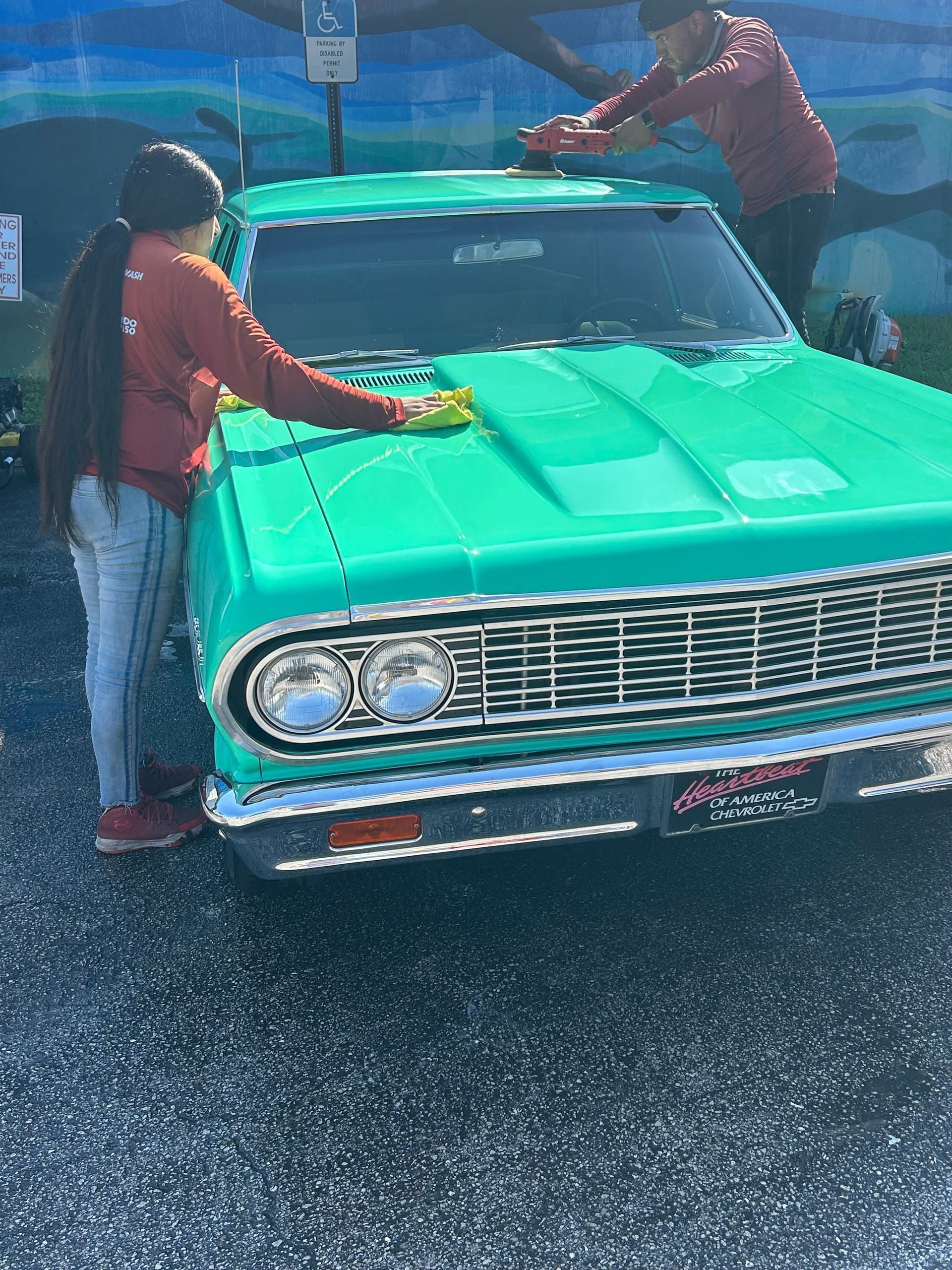 Two people cleaning a bright green classic car. They are wearing red shirts and jeans. Outdoor setting.