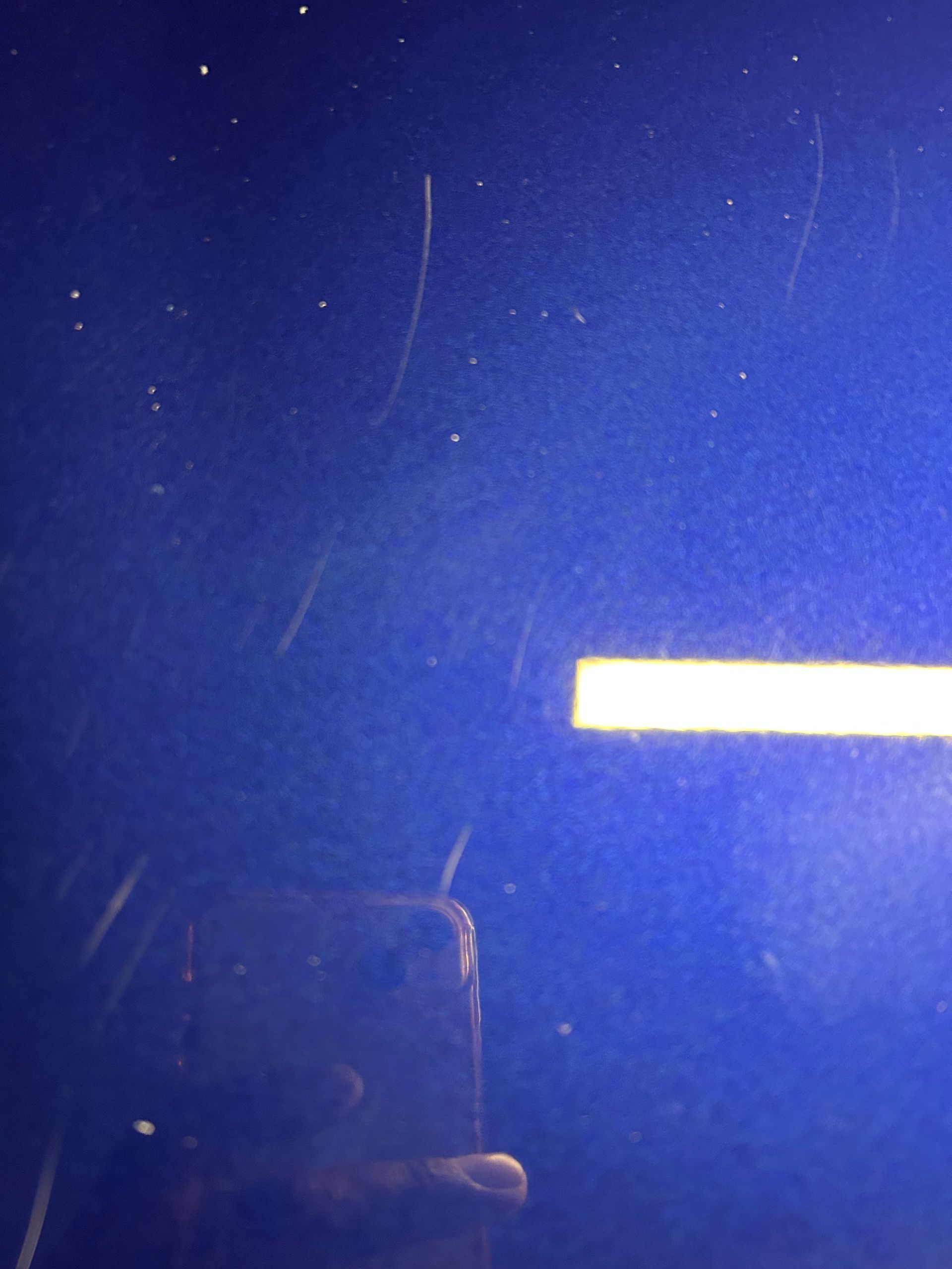 Close-up of a blue car surface with several light scratches and specks. A hand is visible holding a phone in reflection.