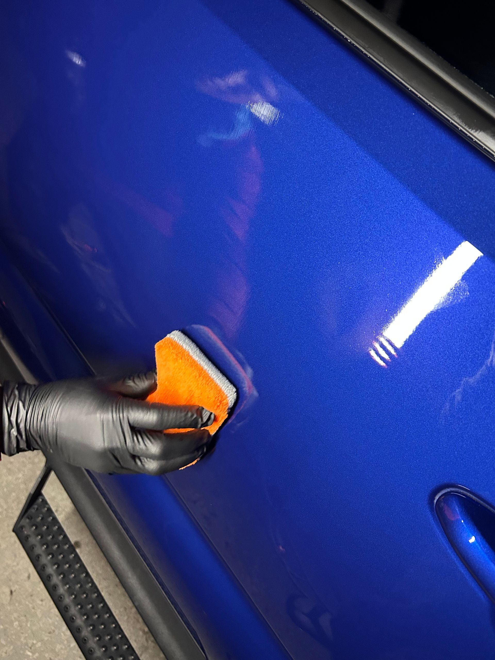 A gloved hand applies detailing product to a blue car door with an orange applicator.