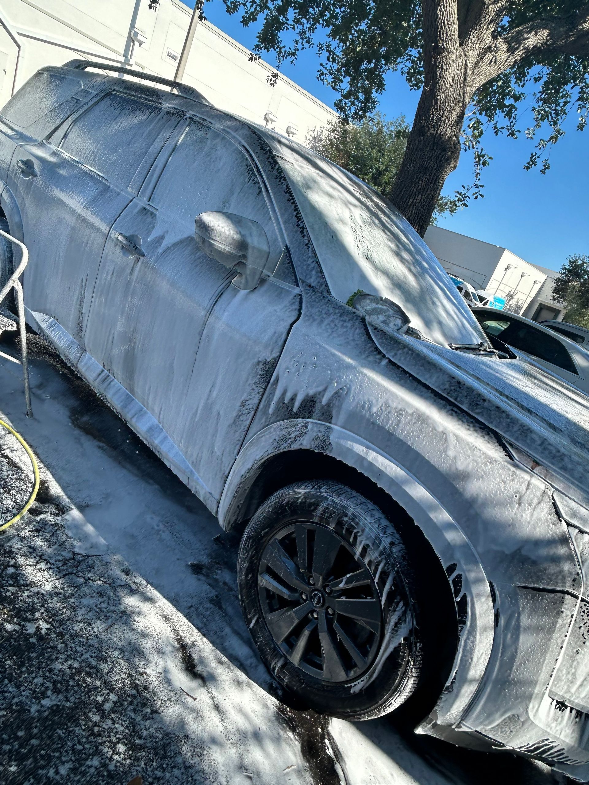 Gray SUV covered in white foam, being washed outdoors with a blue sky background.