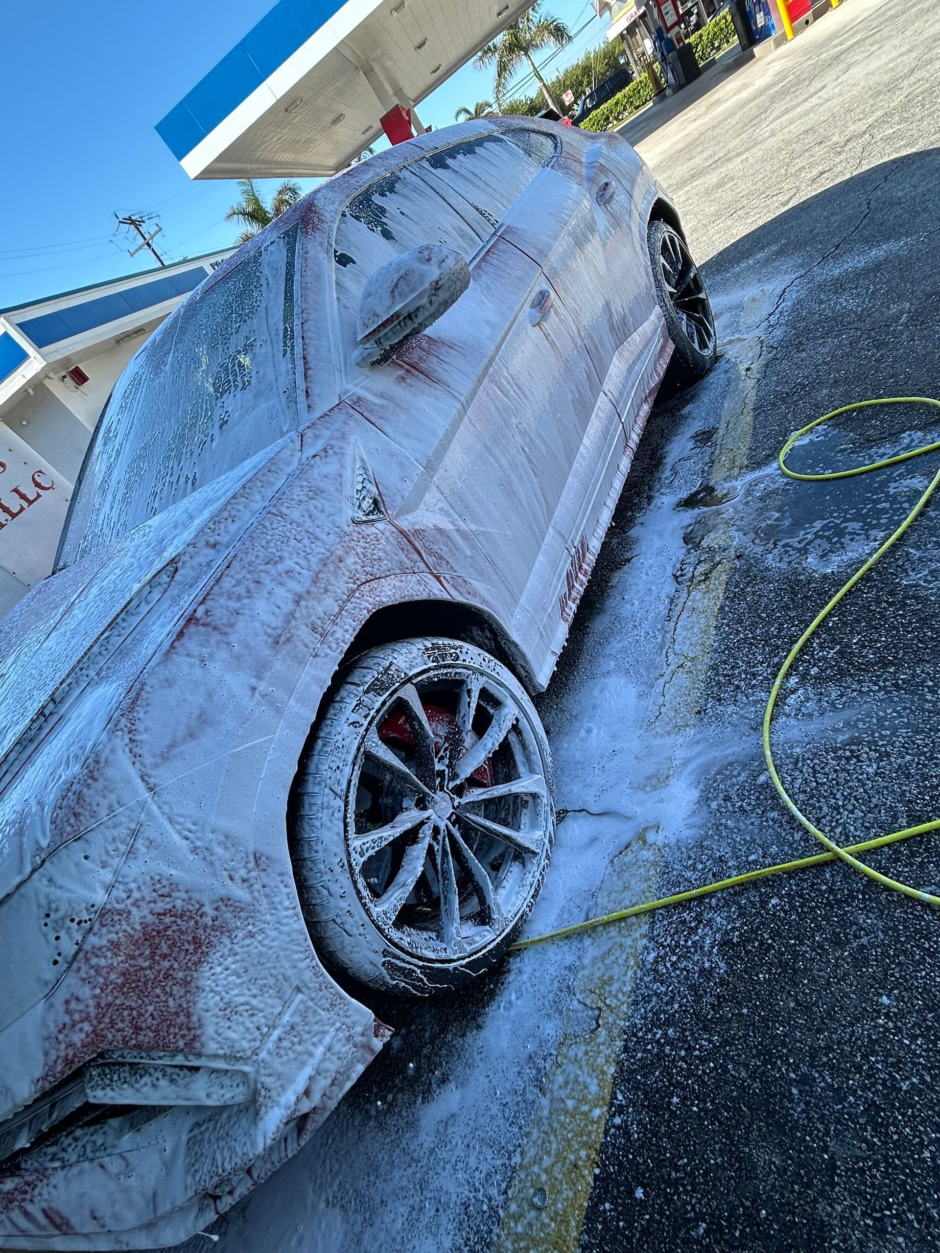 Car covered in white foam at a car wash, red accents visible, yellow hose nearby.