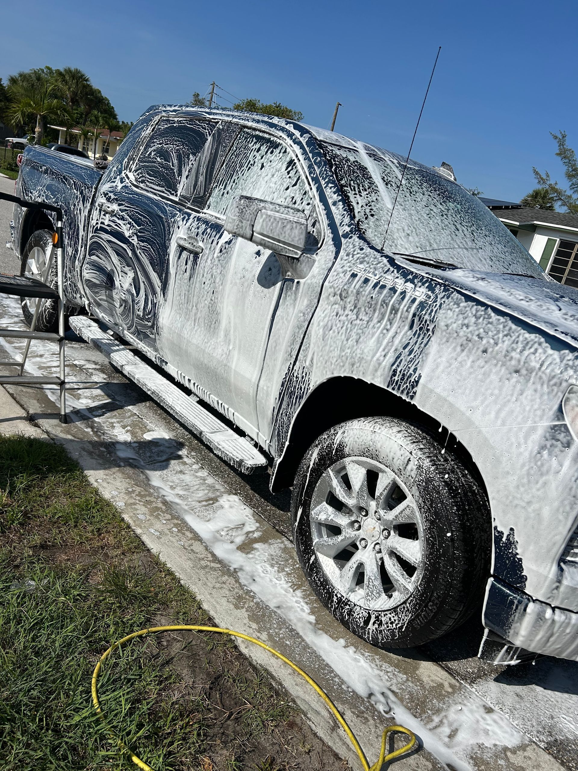 A dark pickup truck covered in white car wash foam sits on a driveway on a sunny day.