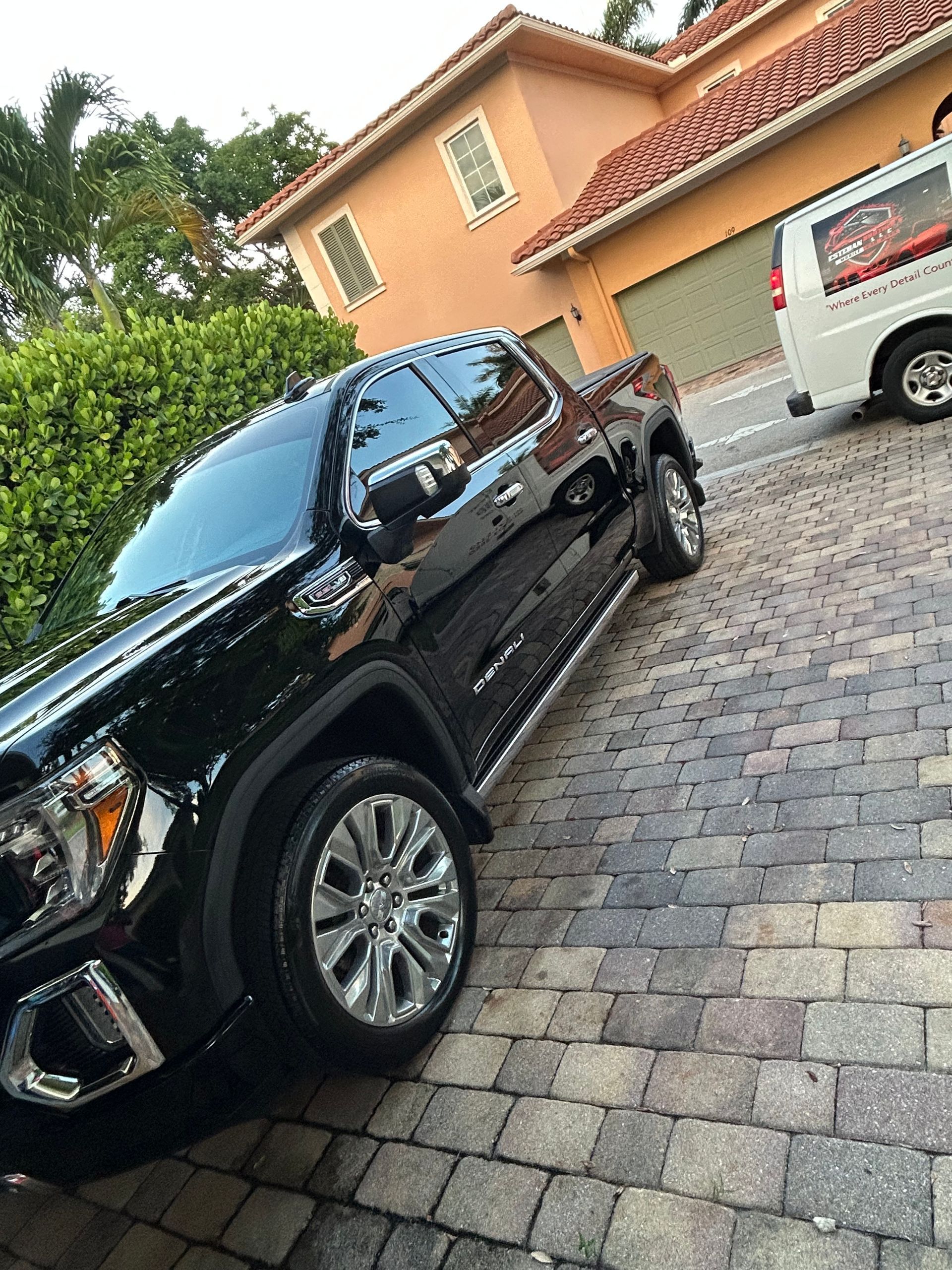 Black GMC truck parked on a brick driveway in front of a house; a white van is parked in the background.