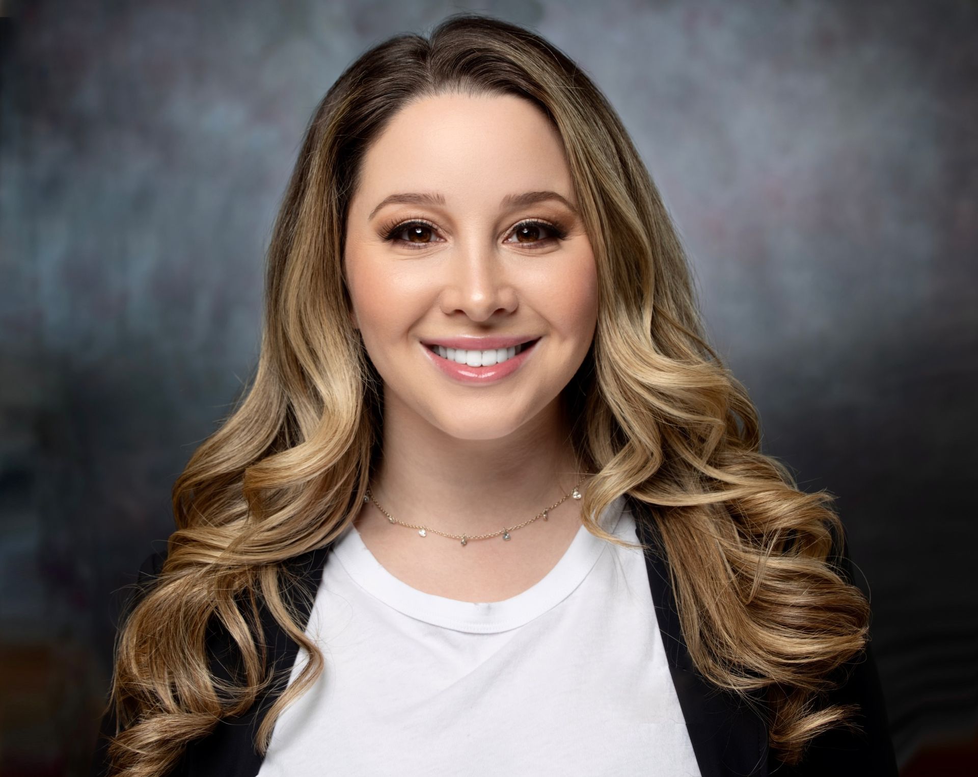 A woman with long hair is smiling for the camera while wearing a white shirt and a black jacket.