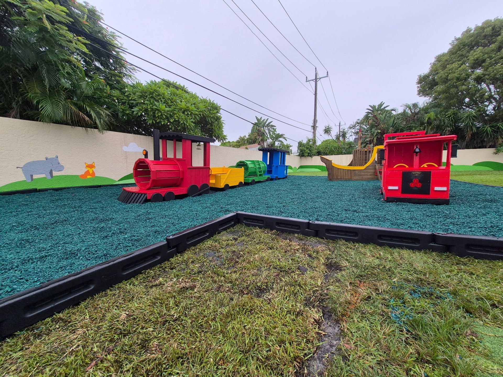 Playground with toy train, grass, and surrounding fence on a cloudy day.