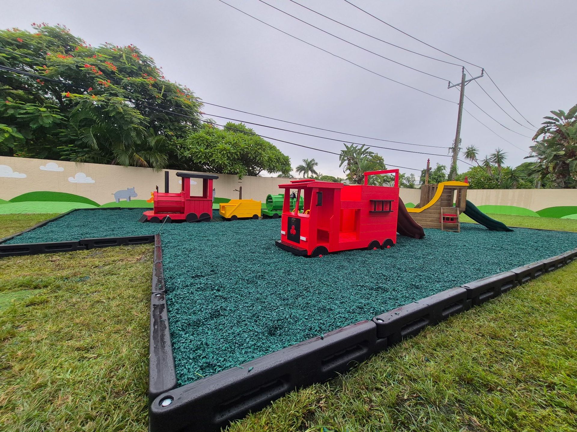 Playground with red train and slide on green rubber surface, surrounded by grass and a wall.