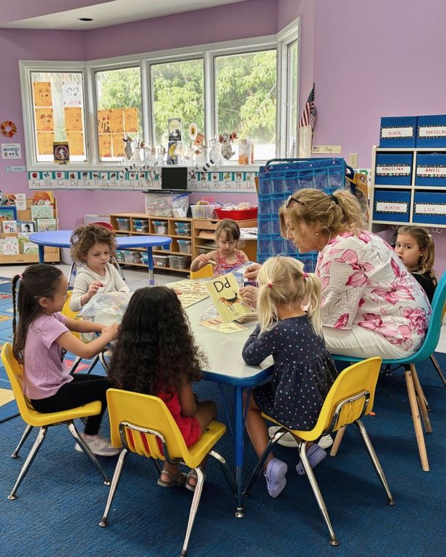 Children and teacher reading book at a table in a classroom.