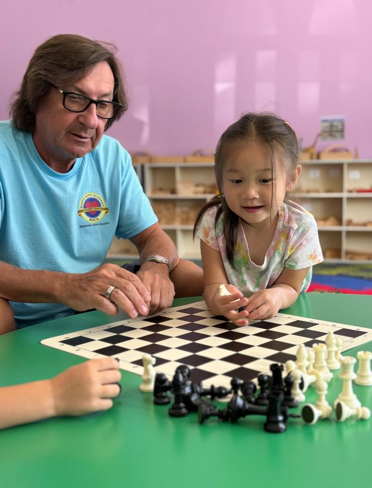 Man and child playing chess, seated at a green table, indoors. The child holds a chess piece, smiling.