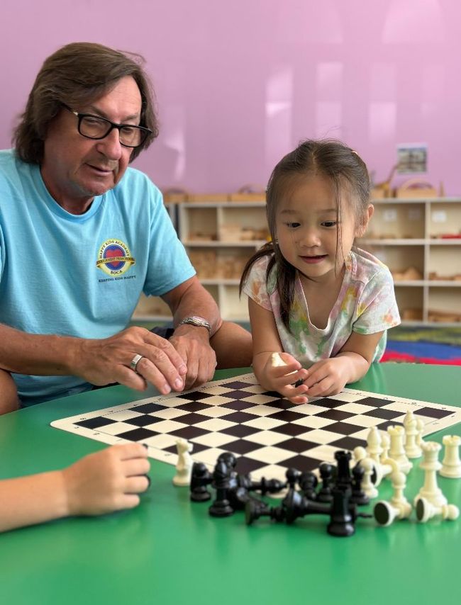Man and child playing chess, seated at a green table, indoors. The child holds a chess piece, smiling.