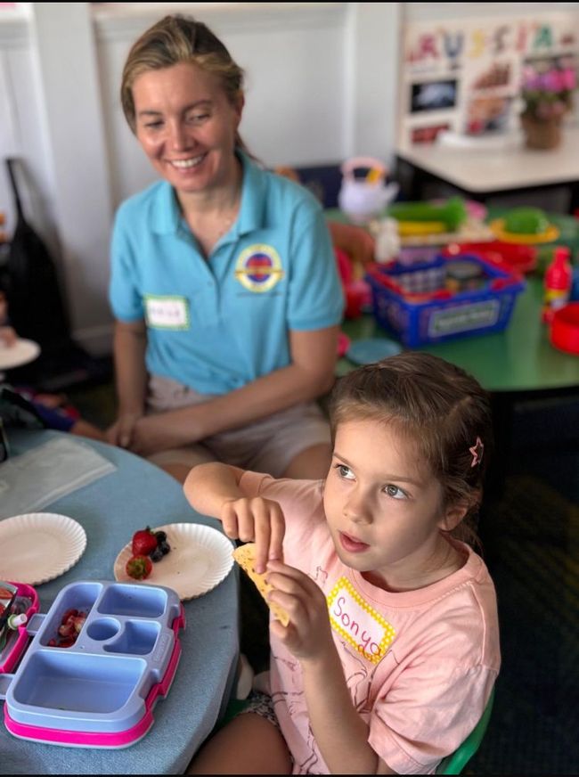 Child eating snack at table with adult; colorful lunchbox, smiling faces, indoors.