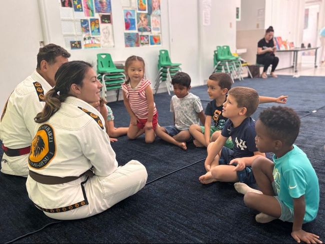A woman in a gi interacts with a group of children, sitting on a blue carpet.