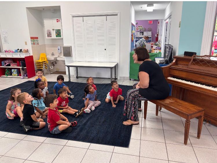 Teacher sits on a bench, facing a group of seated children in a classroom.