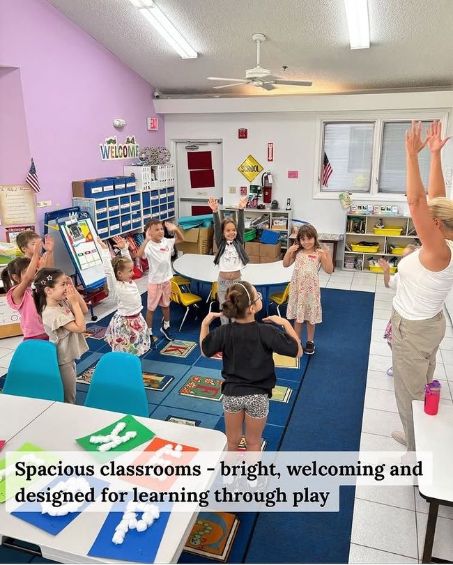 Children and teacher in a bright classroom, hands raised. Purple wall, blue rug, and educational toys.