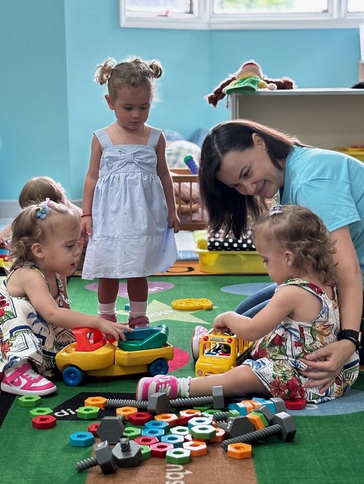 Children and a teacher play with toys on a rug in a classroom.