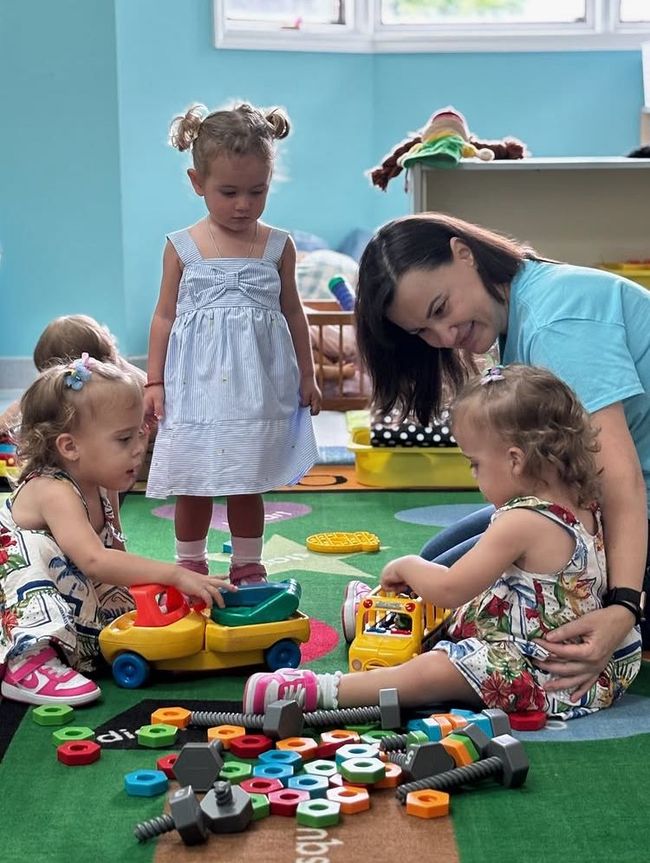 Children and a teacher play with toys on a rug in a classroom.