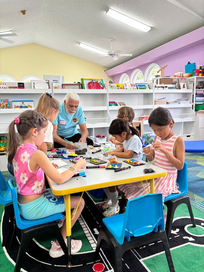 Children and an adult build with blocks at a table in a brightly lit classroom.