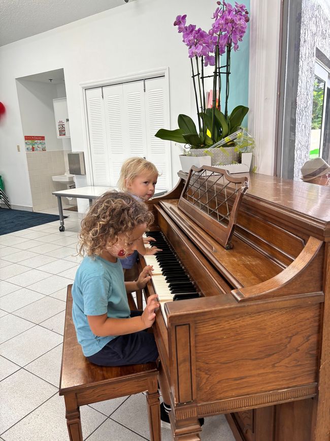 Two children playing a wooden piano indoors, near an orchid plant.
