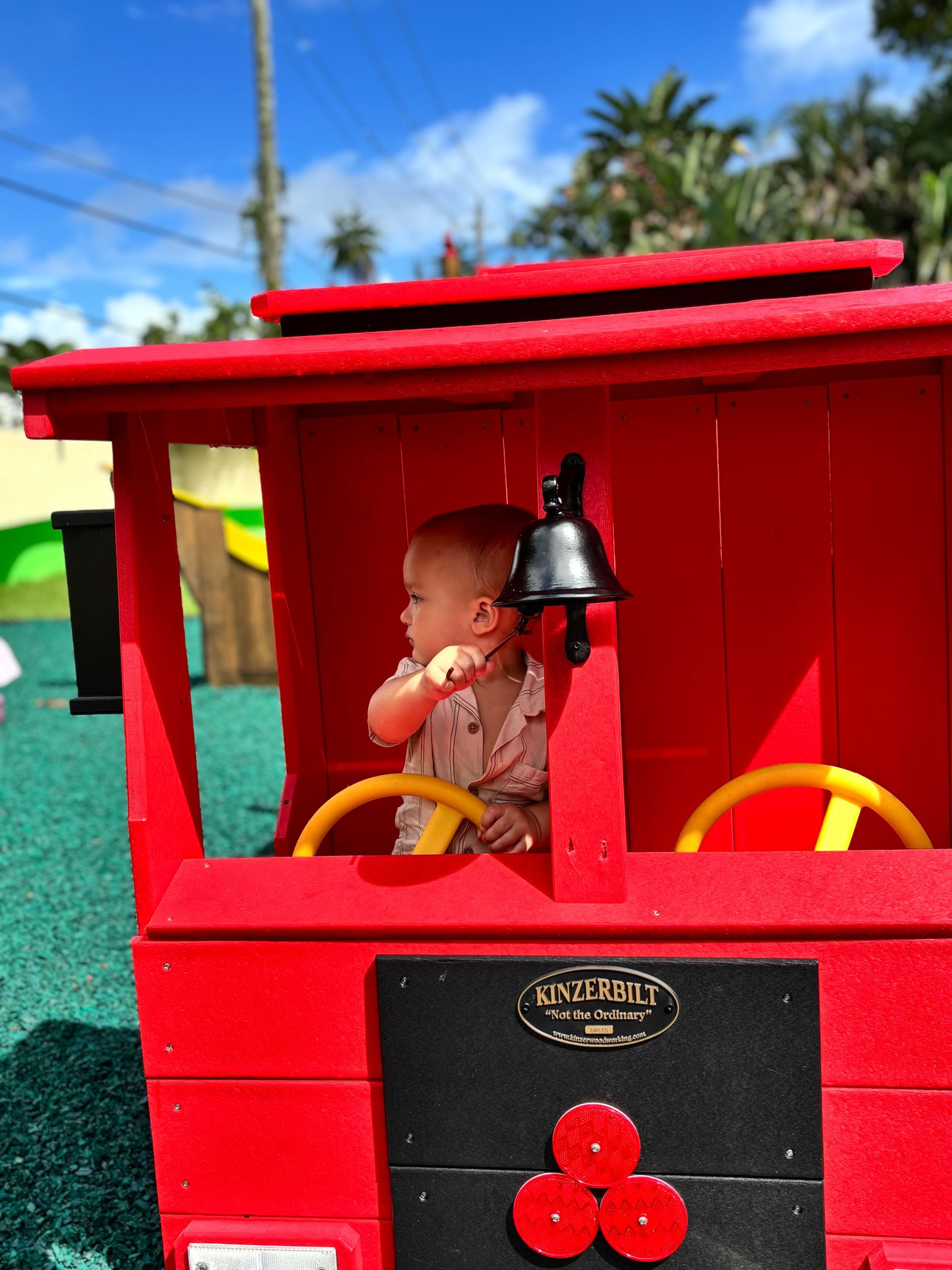 Baby sitting in a red toy train cab, looking to the side. Yellow steering wheels, black bell, outdoor setting.