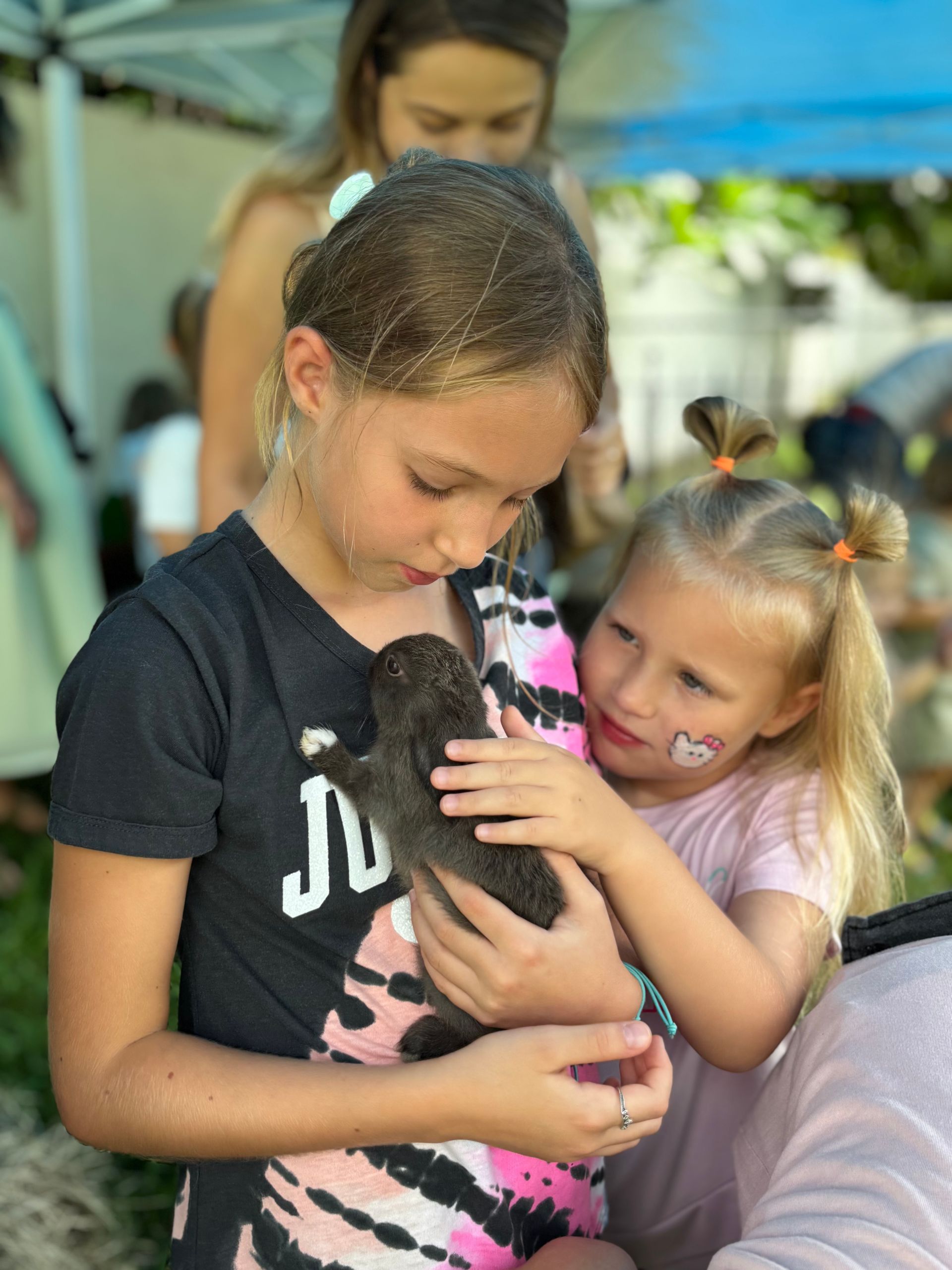 Two girls holding a small dark animal. One girl looks concerned, the other smiles. Outdoors setting.