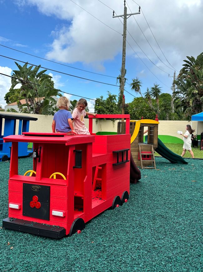 Two children on a red fire truck playset in a playground; a woman walks in background.